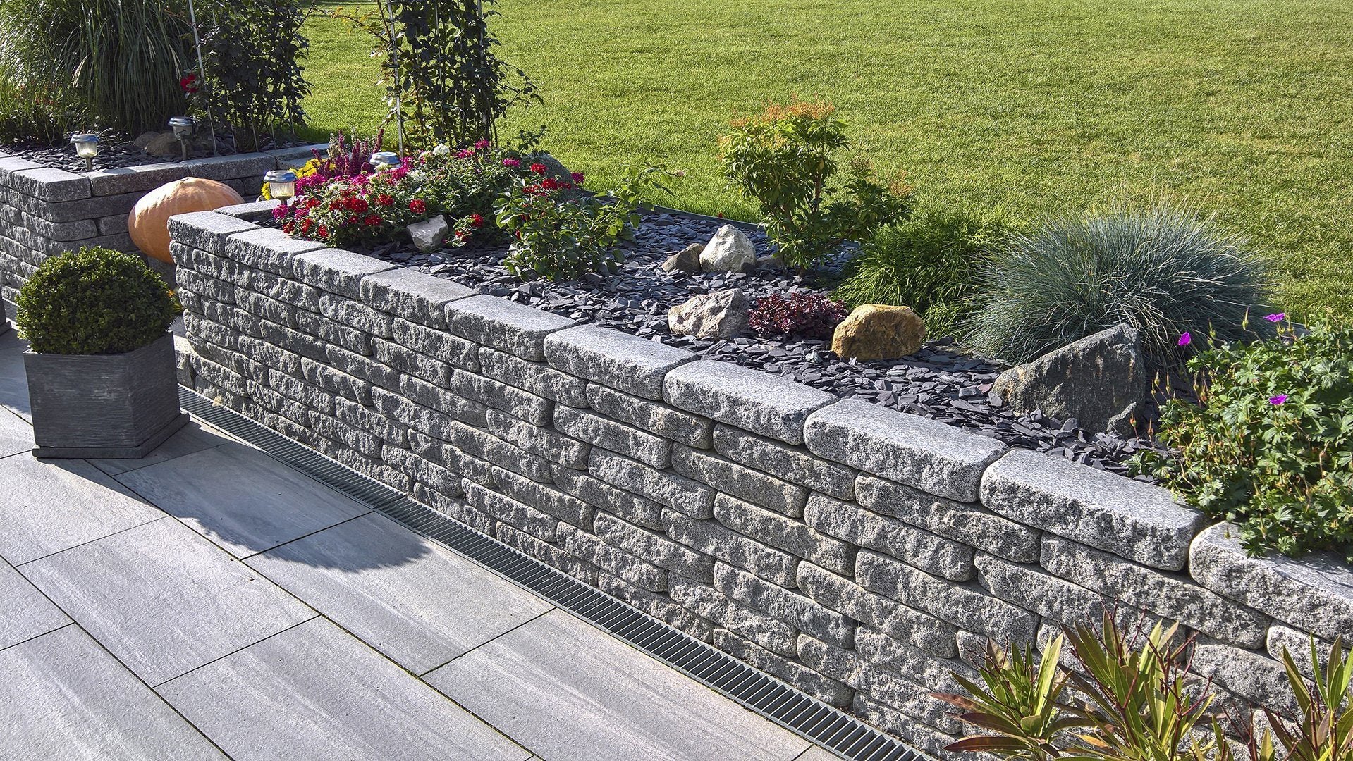 Lage keermuur van grijze stapelblokken met een verhoogde border vol beplanting, naast een terras van grijze tegels